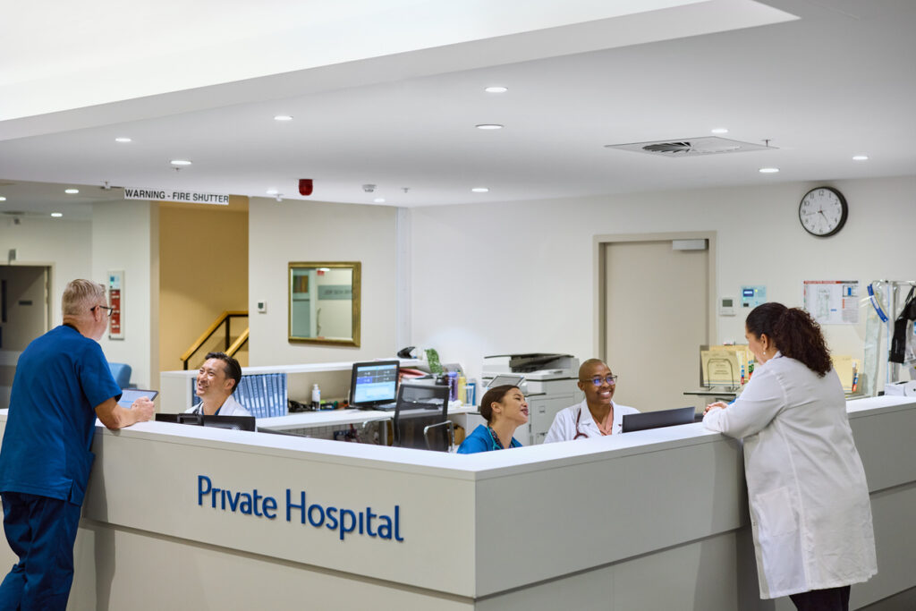 Medical staff interacting at bariatric surgery hospitals reception. Surgeon in blue scrubs using tablet while doctors and nurses smiling and talking. Bright and modern reception area with computers, paperwork, and equipment.
